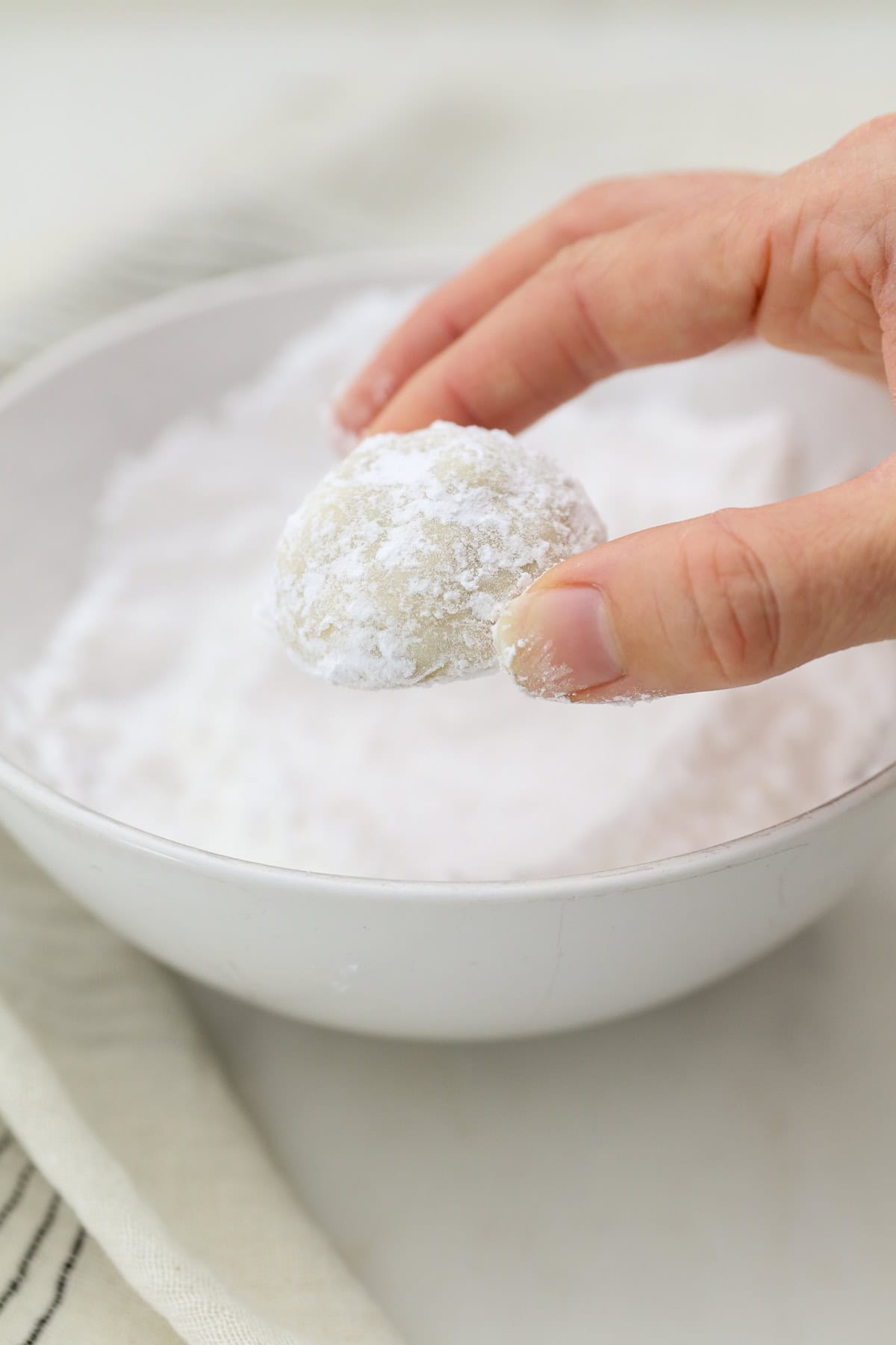 Snowball cookies are easy Christmas cookies made with buttery shortbread and chopped nuts, rolled in powdered sugar to look like snowballs! A hand rolling a snowball cookie in a bowl of powdered sugar.