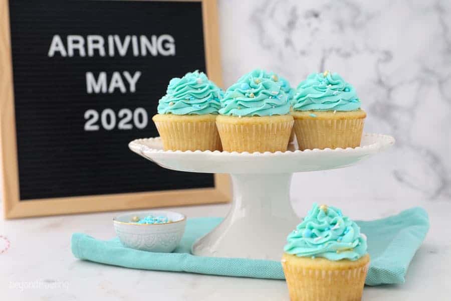 A letter board that says "arriving May 2020" with some blue frosted cupcakes