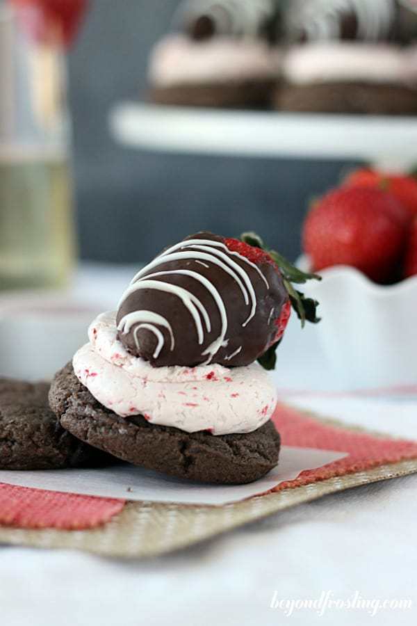 A chocolate cookie with strawberry frosting and a chocolate covered strawberry on top, a cup of strawberries in the background