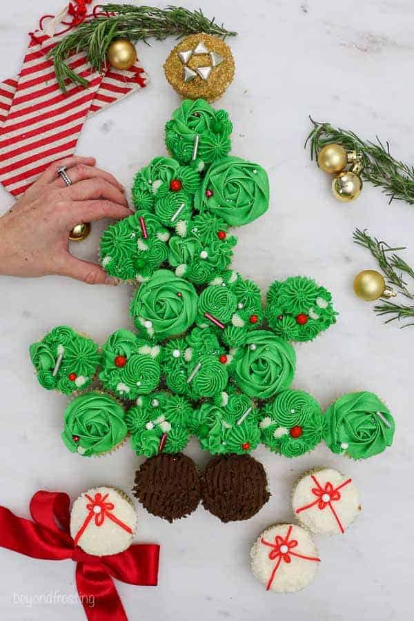 A hand grabbing a cupcake from a Christmas tree cake on a countertop with stockings and other holiday decorations