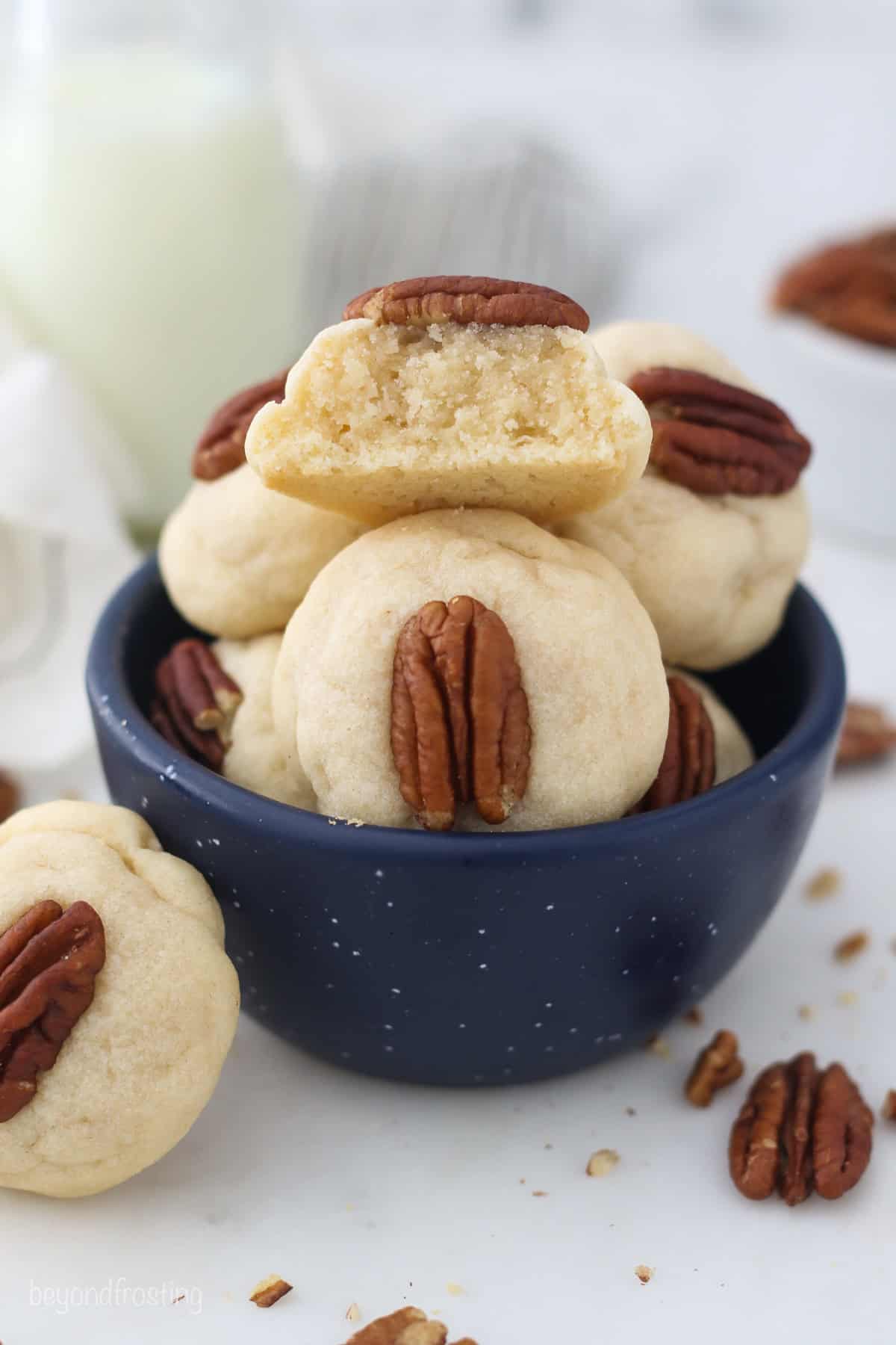 A cream cheese cookie garnished with a pecan leaning against a bowl of cream cheese cookies, with a bite missing from one of the cookies.