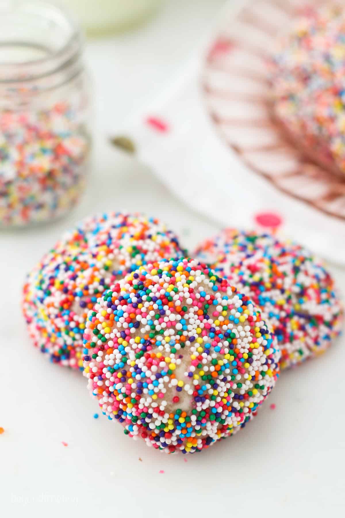 Three sprinkle-covered cream cheese cookies in a small pile, with a jar of colorful sprinkles in the background.