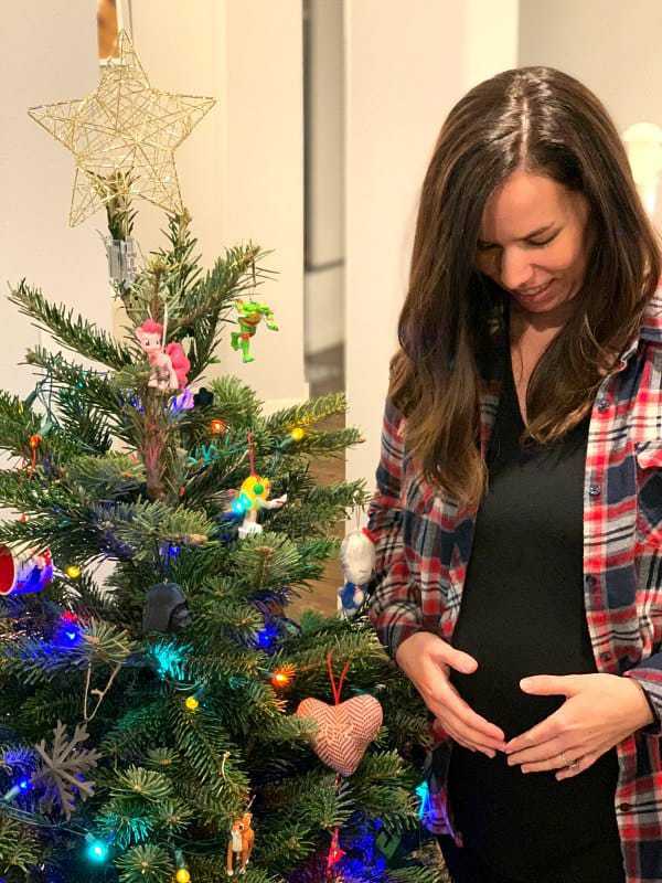 A pregnant woman looking down at her stomach and standing in front of a Christmas tree