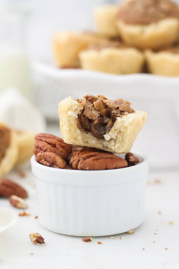 A small white dish filled with pecans with a mini pecan tart on top, with a bite taken out of it to show the gooey filling