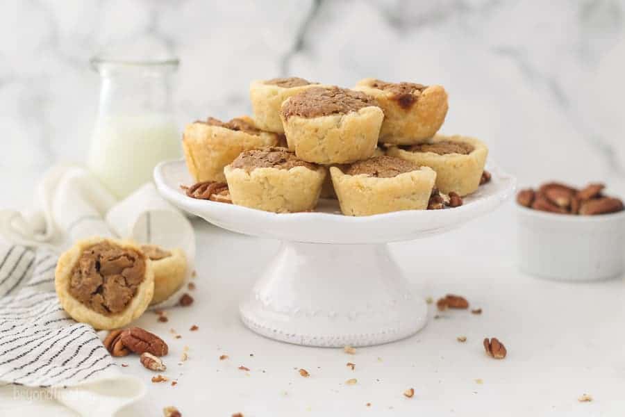 A wide angle shot of pecan tarts on a small white cake stand, with a jar of milk in the background