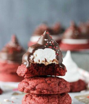 A stack of 3 red velvet cookies, the one on top is frosted and coated in chocolate, has a bite missing