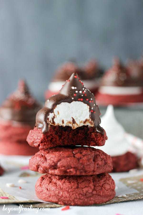 A stack of 3 red velvet cookies, the one on top is frosted and coated in chocolate, has a bite missing