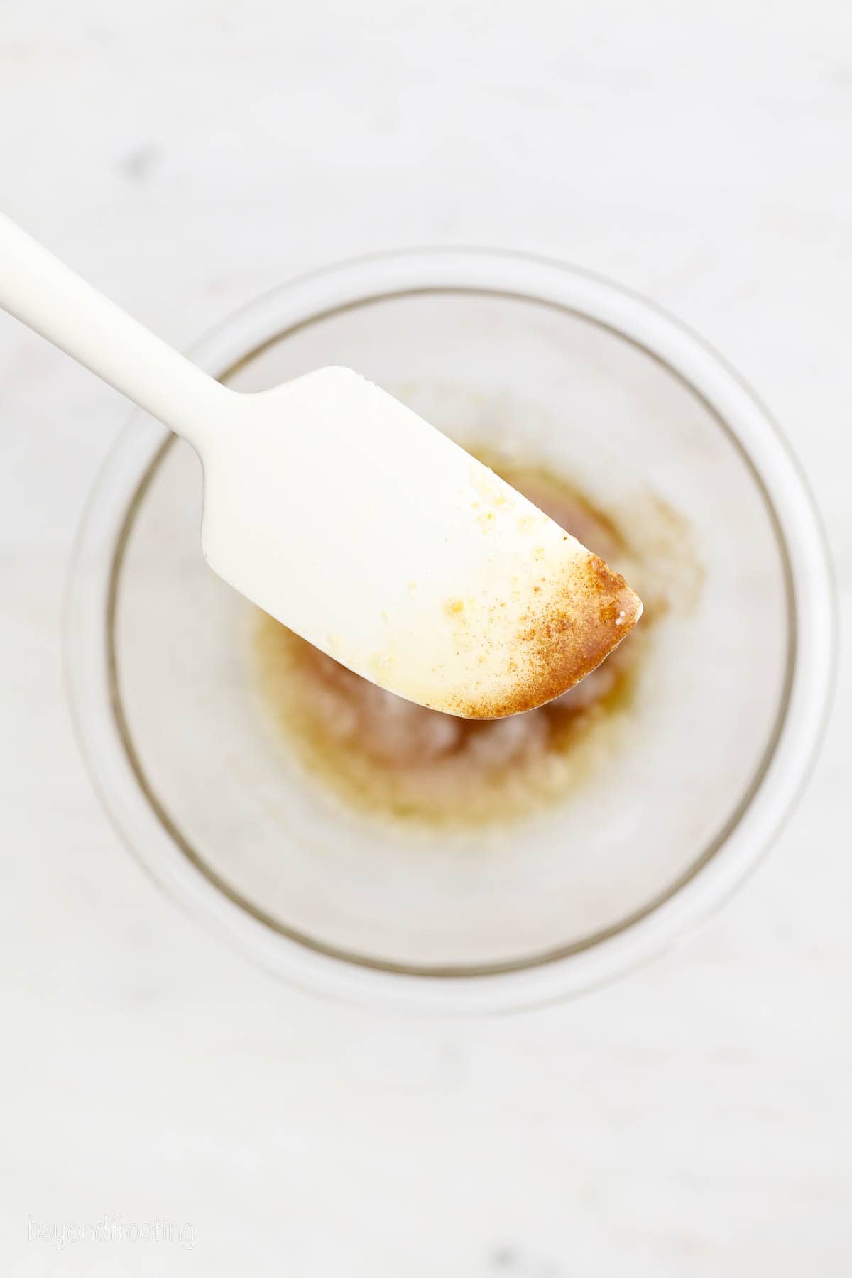Close up of a white rubber spatula held up to show brown flecks of toasted milk solids over a bowl of brown butter.