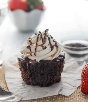 An unwrapped chocolate cupcake on a piece of burlap with a bowl of strawberries in the background