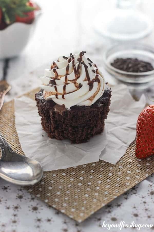 An unwrapped chocolate cupcake on a piece of burlap with a strawberry and a spoon next to it