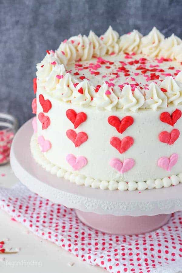 A layer cake with vanilla frosting decorated with 3 shades of red and pink buttercream hearts sitting on a pink cake stand and a red polka dot napkin