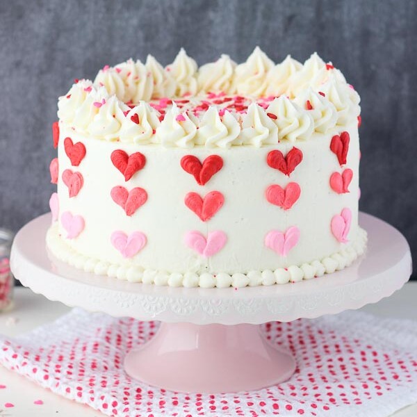 A layer cake with vanilla frosting decorated with 3 shades of red and pink buttercream hearts sitting on a pink cake stand and a red polka dot napkin
