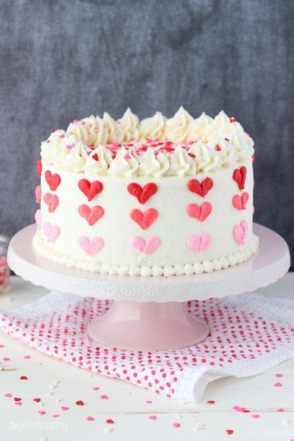 A layer cake with vanilla frosting decorated with 3 shades of red and pink buttercream hearts sitting on a pink cake stand and a red polka dot napkin