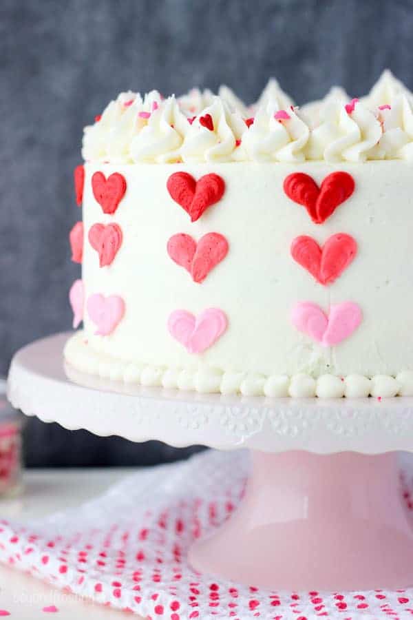 A close up shot of a layer cake with vanilla frosting decorated with 3 shades of red and pink buttercream hearts sitting on a pink cake stand