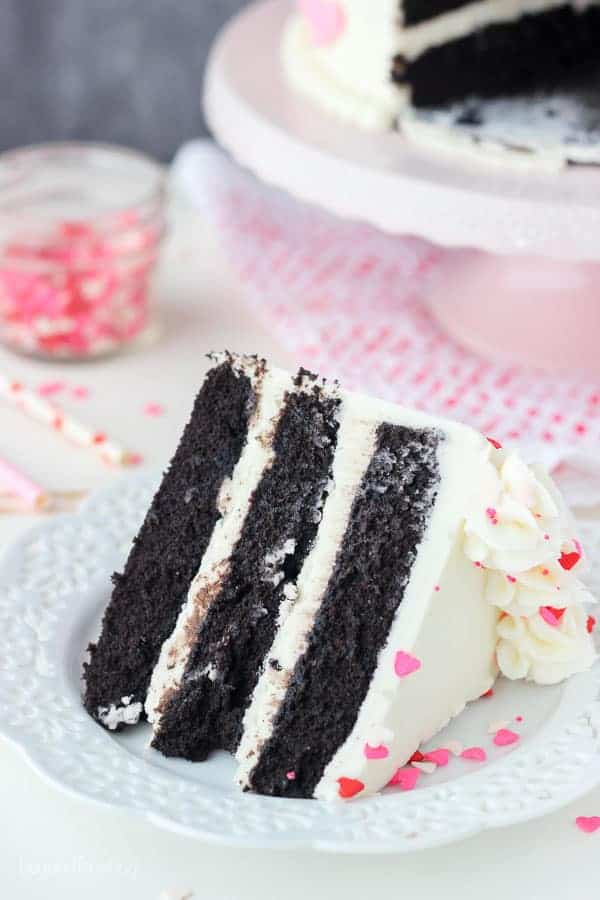 A 3 layer slice of chocolate cake with vanilla frosting on a white plate sprinkled with heart shaped sprinkles