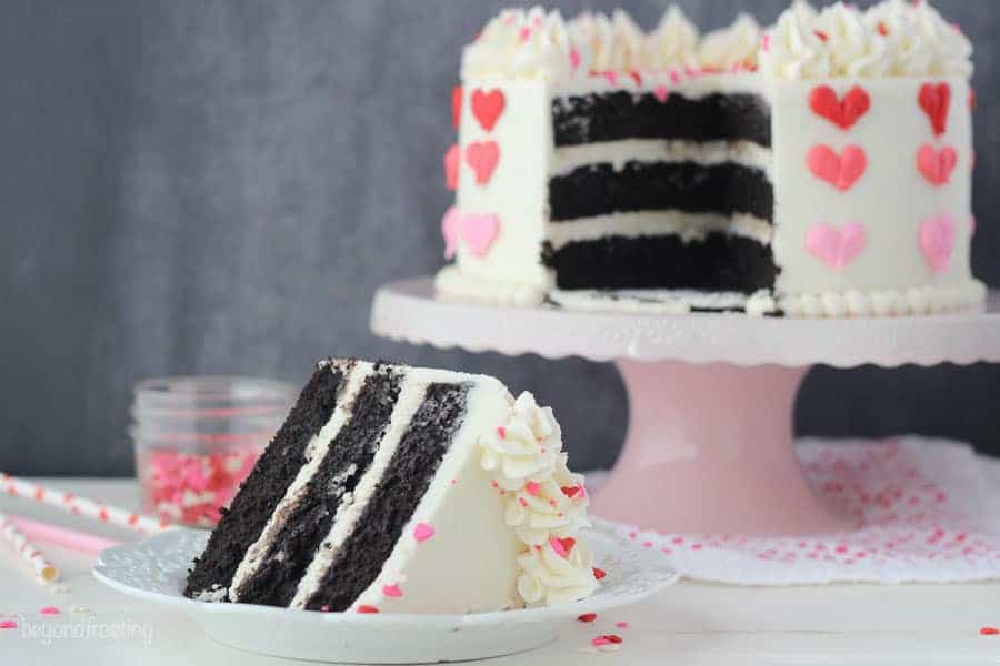 A slice of chocolate layer cake, a pink cake stand in the background with the cake, missing a slice
