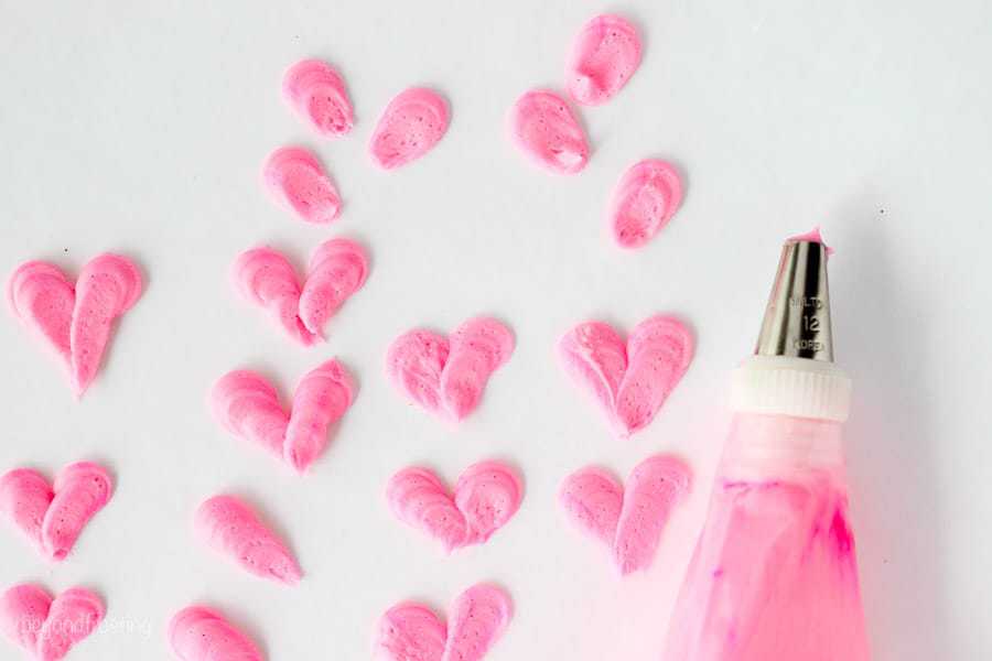 Pink buttercream hearts piped on a piece of parchment paper with a piping bag sitting next to it