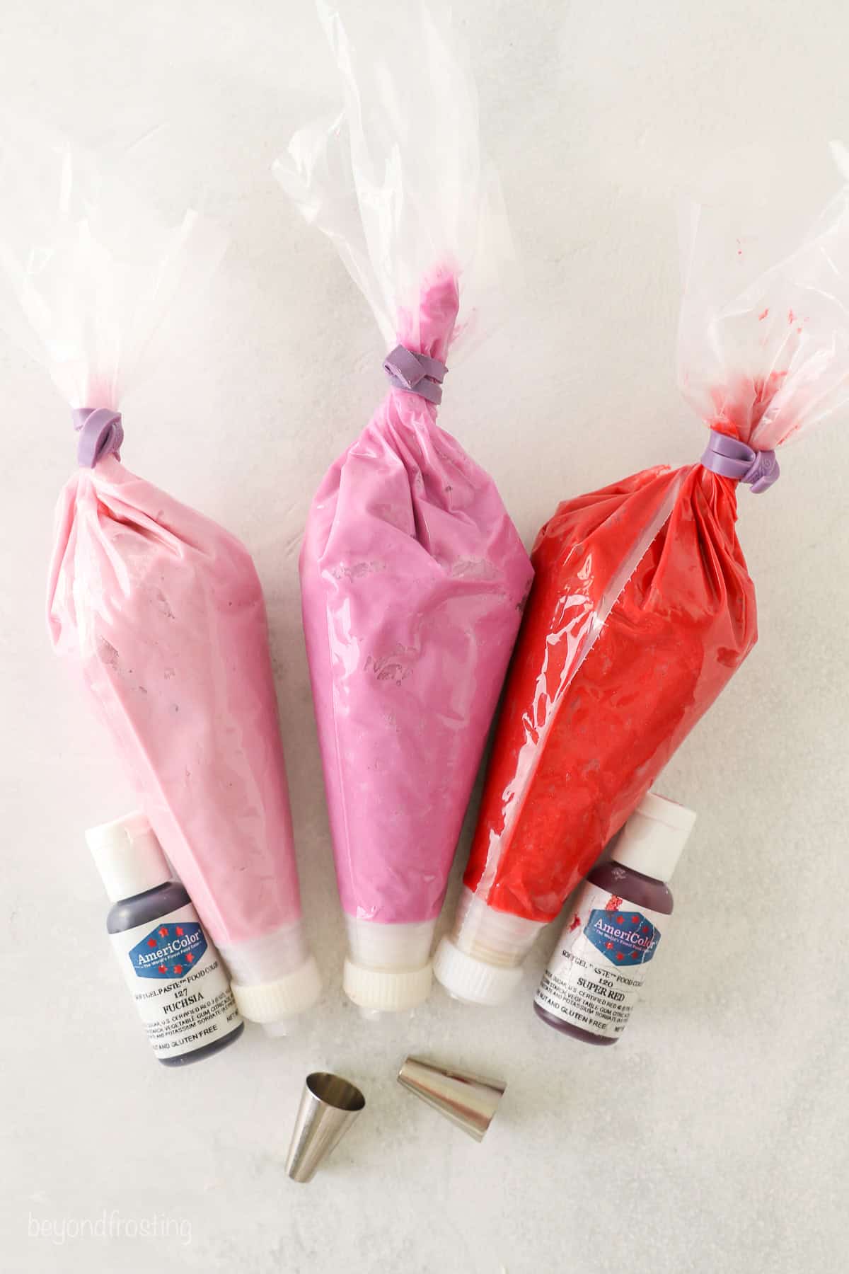 Overhead view of three piping bags filled with light pink, dark pink, and red frosting next to two small bottles of gel food coloring.