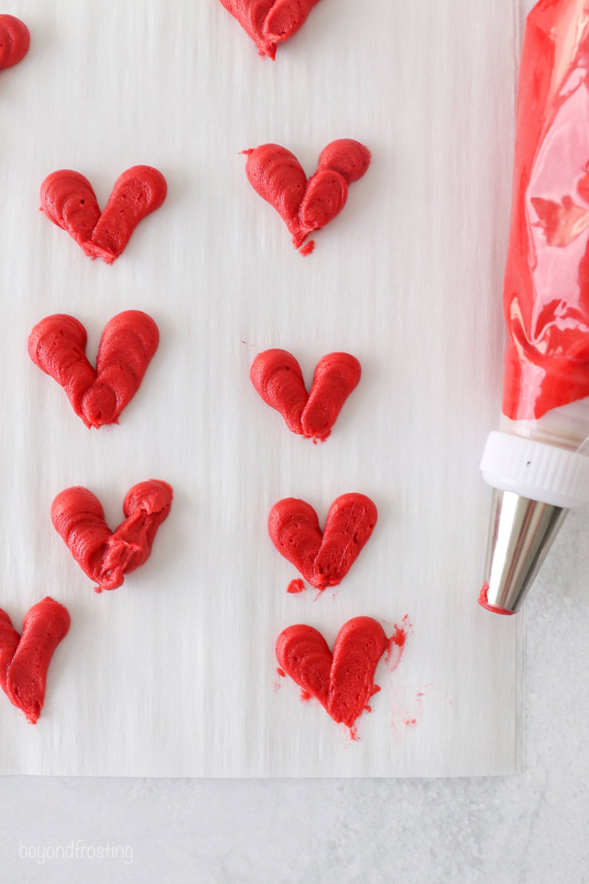 Overhead view of a piping bag with red frosting laying nest to piped red frosting hearts on a sheet of parchment paper.