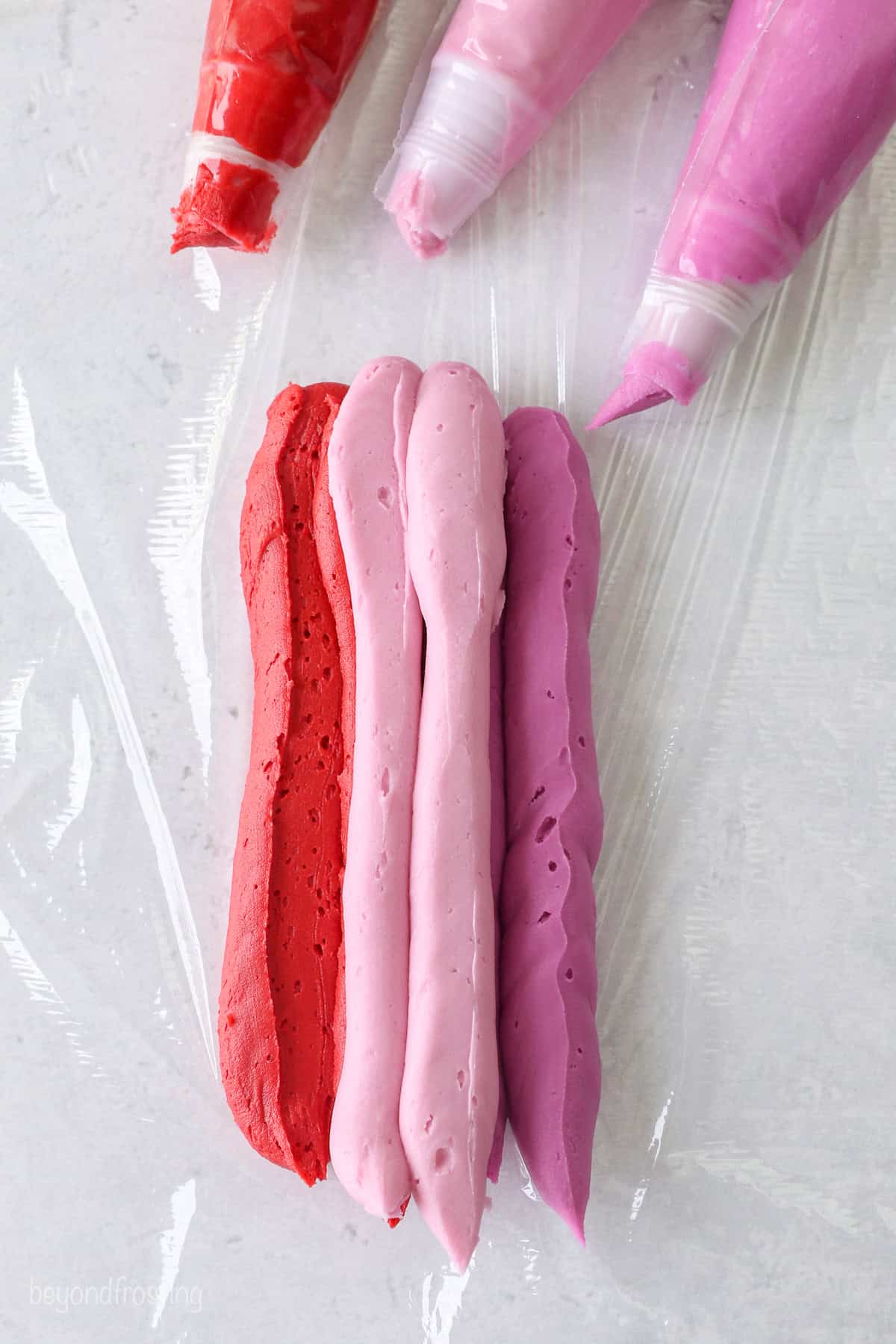 Piping bags laying next to piped frosting in varying shades of red and pink on a piece of plastic wrap.