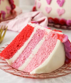 A slice of Valentine's Day ombre cake on a plate with the rest of the cake in the background.