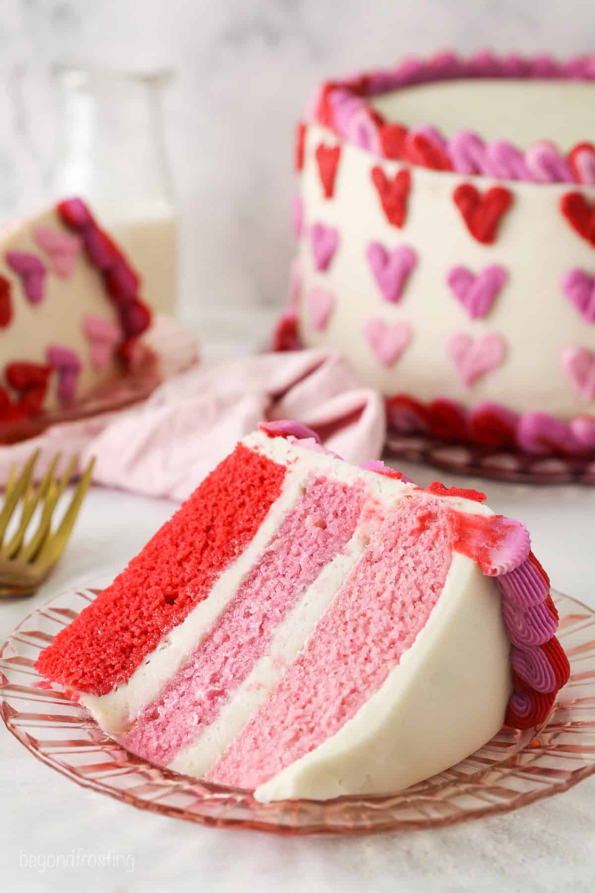 A slice of Valentine's Day ombre cake on a plate with the rest of the cake in the background.