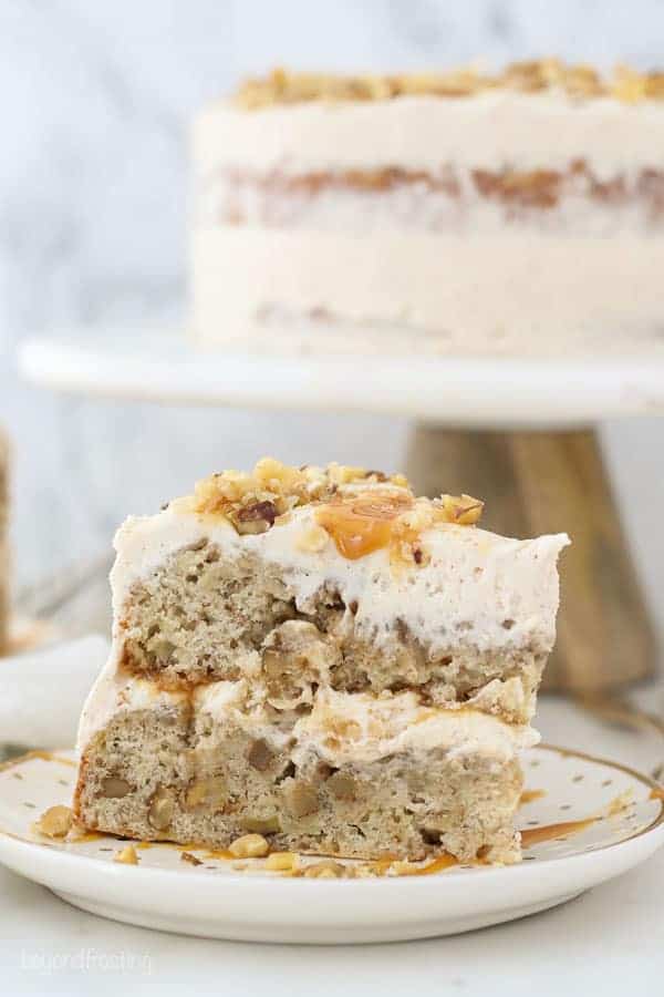 An side view of a slice of banana cake and a cake stand blurred out in the background with frosting and caramel dripping down the side