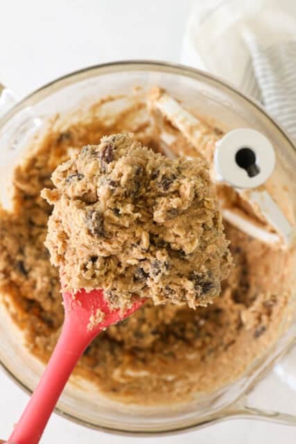 Overhead view of a red rubber spatula scooping oatmeal raisin cookie dough from a mixer bowl.