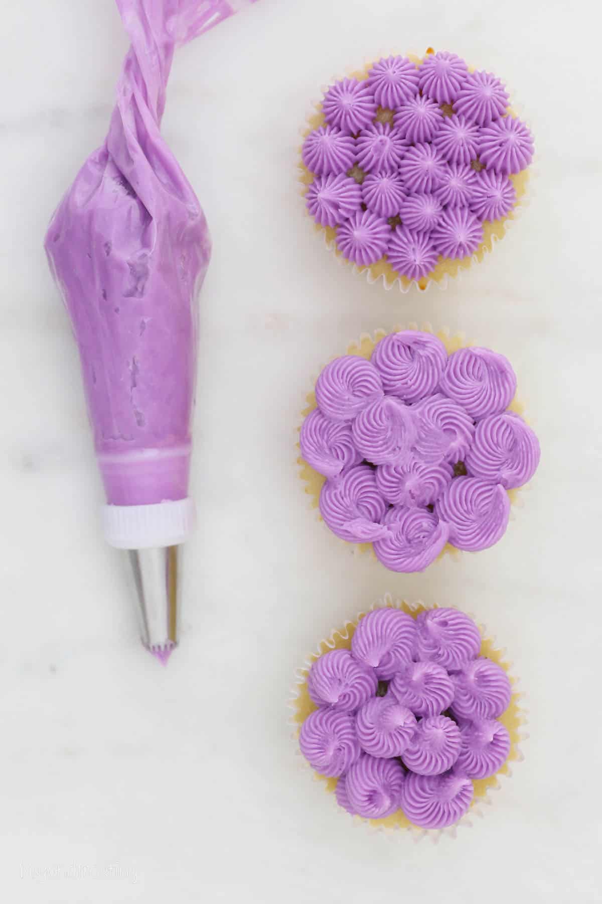 Three cupcakes decorated with purple frosting using various French star piping tips, next to a piping bag of purple buttercream frosting.