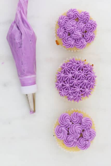 Three cupcakes decorated with purple frosting using various open star piping tips, next to a piping bag of purple buttercream frosting.