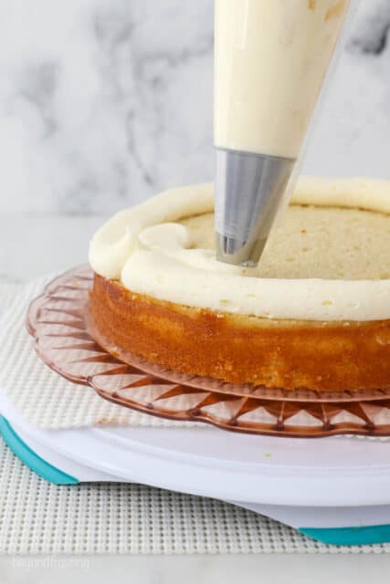 A piping bag pipes a layer of vanilla frosting over the first layer of cake on a plate.