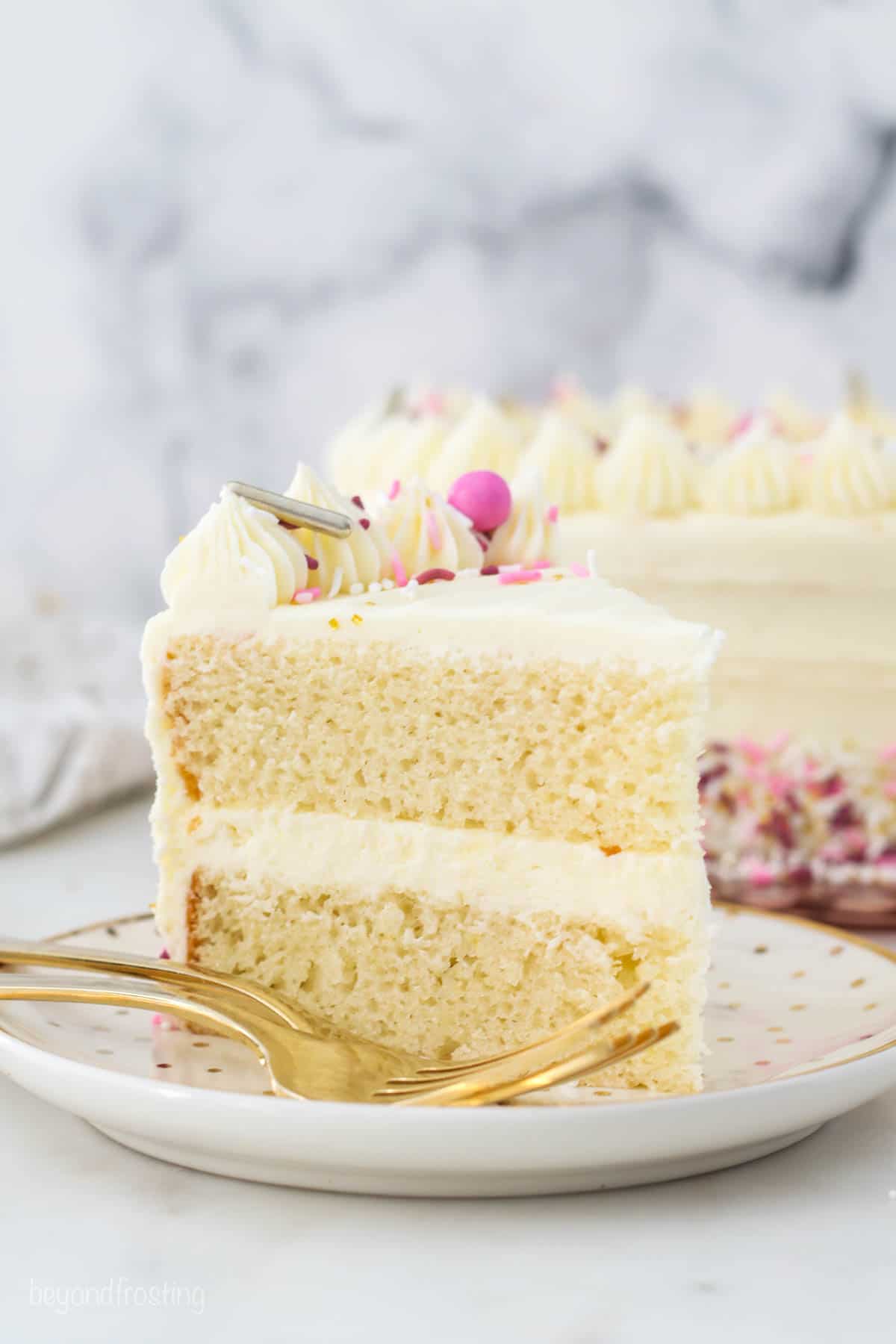 A slice of frosted layer cake decorated with frosting swirls and rainbow sprinkles on a white plate next to a fork.