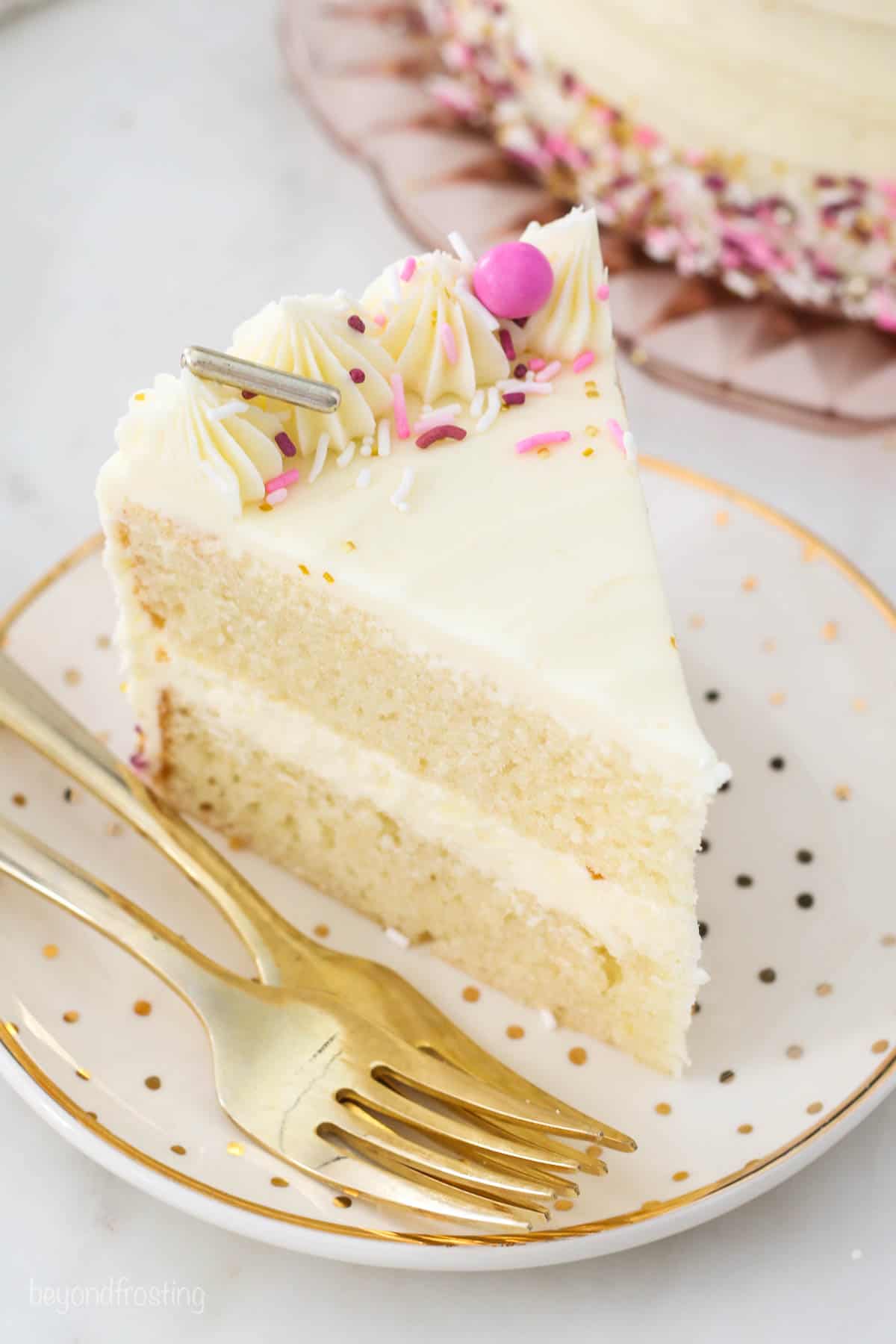 A slice of frosted layer cake decorated with frosting swirls and rainbow sprinkles on a white plate next to a fork.