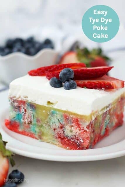 A slice of vanilla poke cake with tye dye colors topped with whipped cream and berries and a text overly