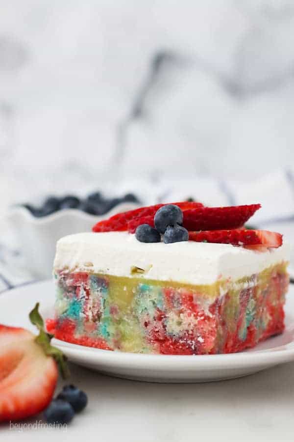 A slice of vanilla poke cake with tye dye colors topped with whipped cream and berries