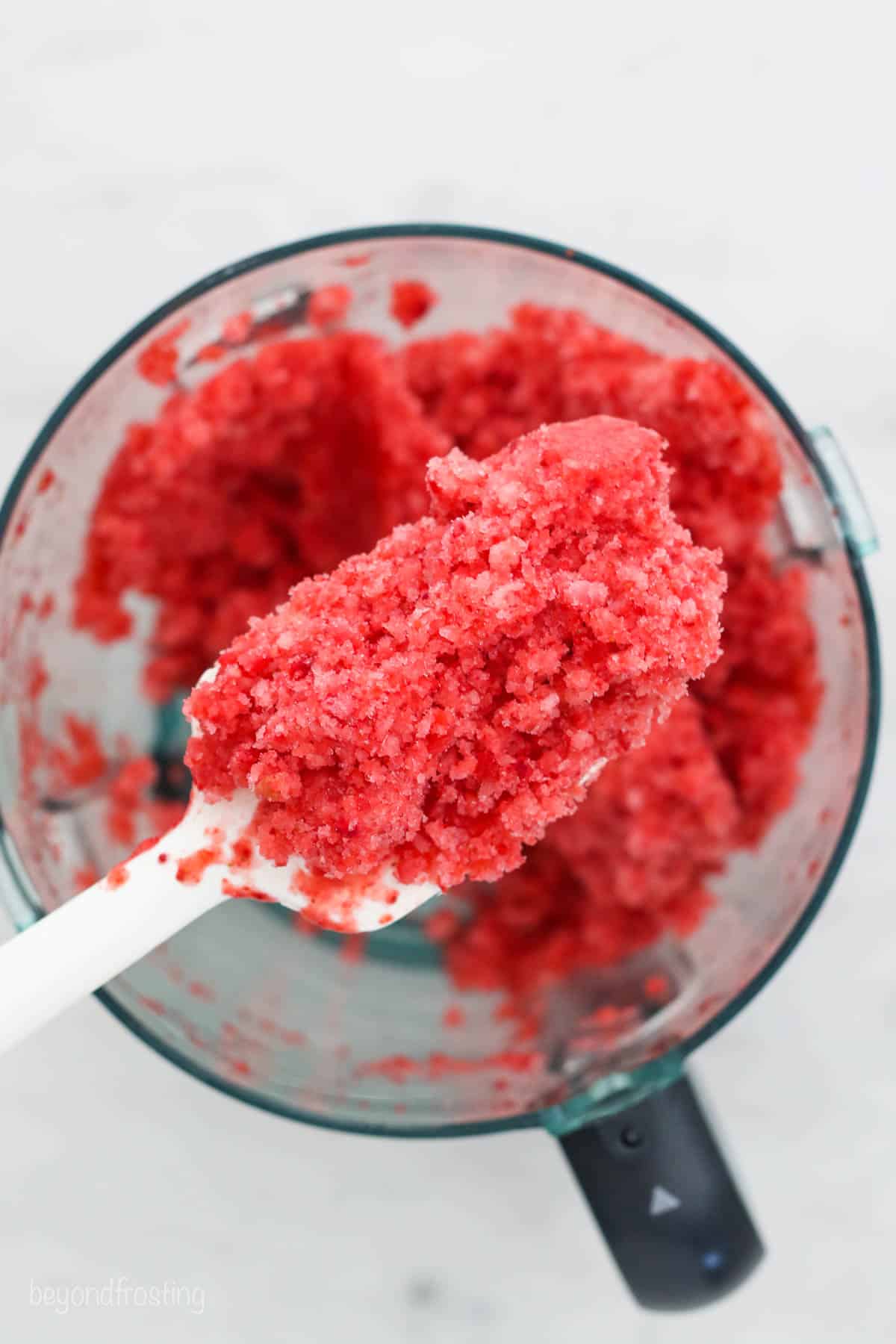 Overhead view of a white spatula lifting a scoop of pureed frozen strawberries from a blender.