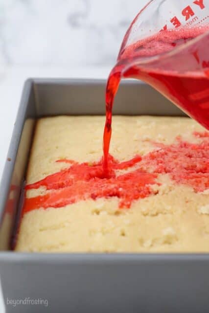 A measuring cup pouring red jello over a vanilla cake
