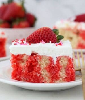 Side view of a slice of poke cake on a plate, showing the red jello on the inside, garnished with a strawberry.