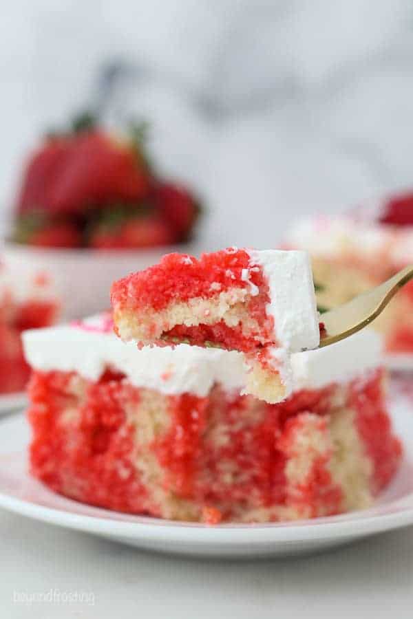 A forkful of strawberry jello poke cake held in front of a slice of cake on a white plate.