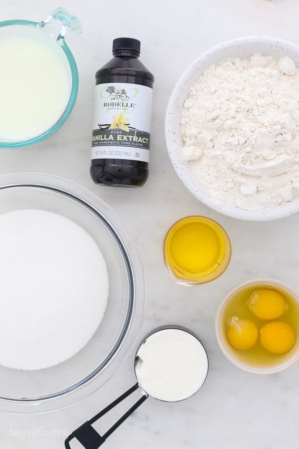 an overhead shot of ingredients for vanilla cupcakes: milk, sugar, flour, eggs, sour cream, oil, vanilla