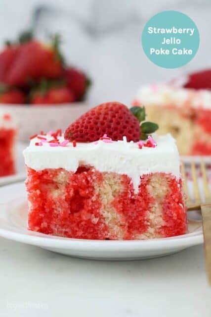 A slice of strawberry jello poke cake garnished with a fresh strawberry on a white plate.