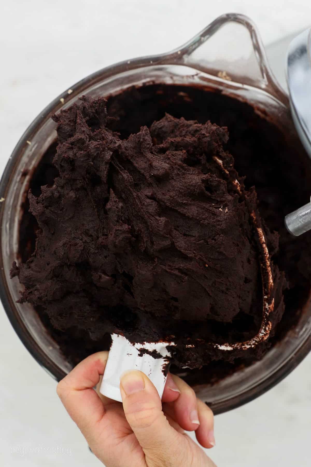 A hand holding a stand mixer attachment covered with chocolate cookie dough above a mixing bowl.