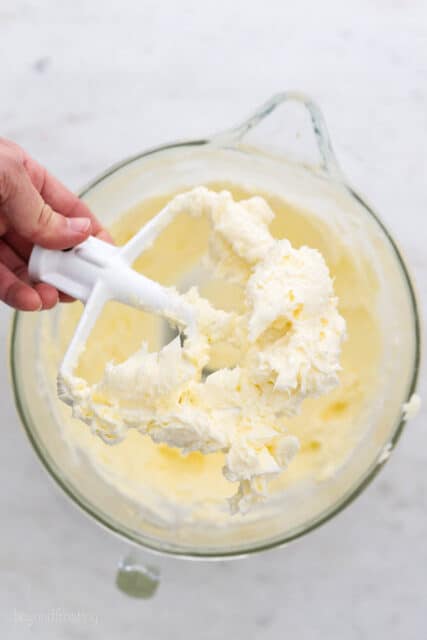 A hand holding a stand mixer whisk attachment over a bowl of frosting.