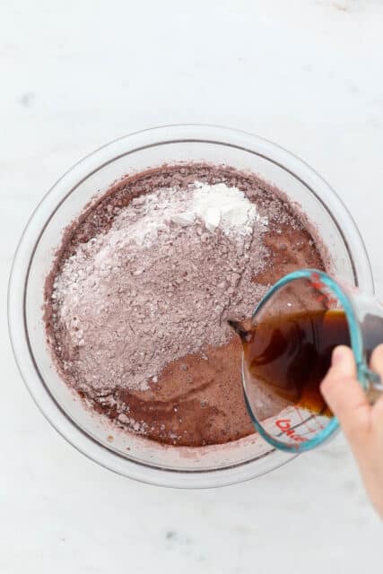 Brewed coffee poured over cocoa powder and dry ingredients added to chocolate cake batter in a glass bowl.