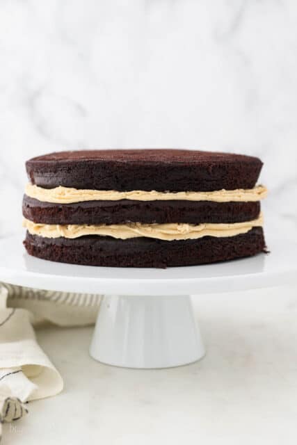 Three layers of chocolate cake with peanut butter frosting on a white cake stand.