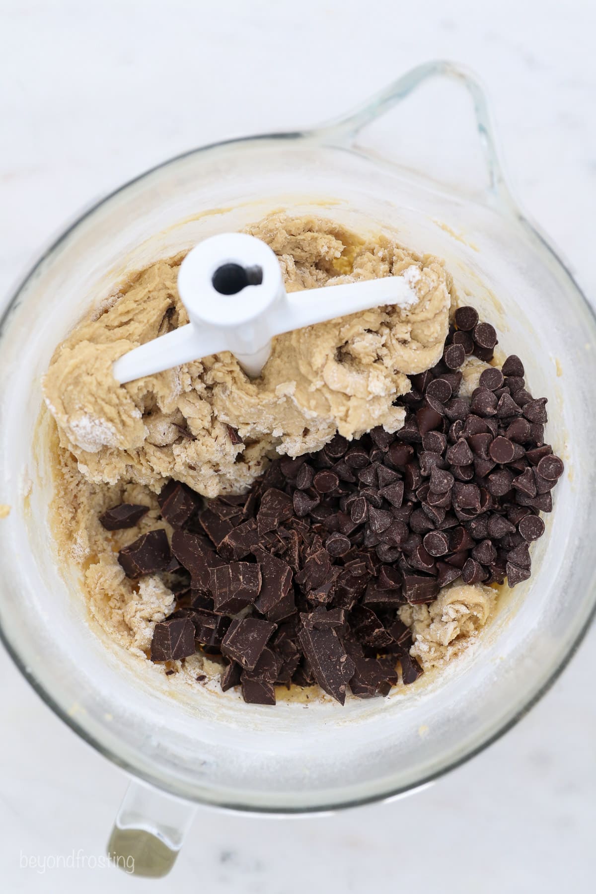 A stand mixer attachment resting in a glass bowl with chocolate chips added to cookie dough.