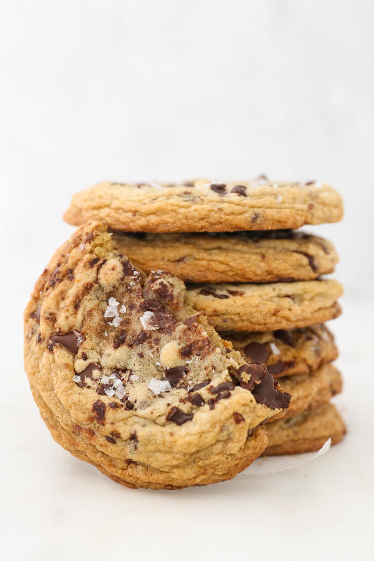One half of a giant chocolate chip cookie leaning against a stack of cookies on a white surface.