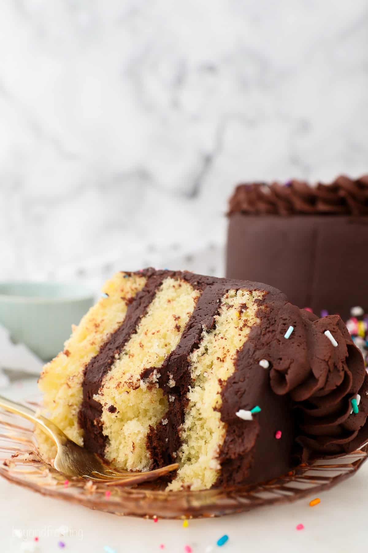 A partially eaten slice of yellow layer cake with chocolate frosting on a plate, with the rest of the cake in the background.