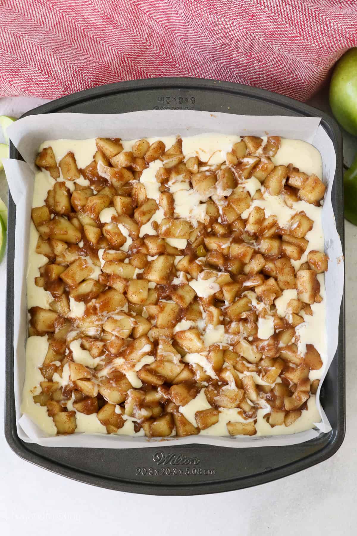 Overhead view of apple pie filling added over top of the cheesecake batter in a lined square baking pan.