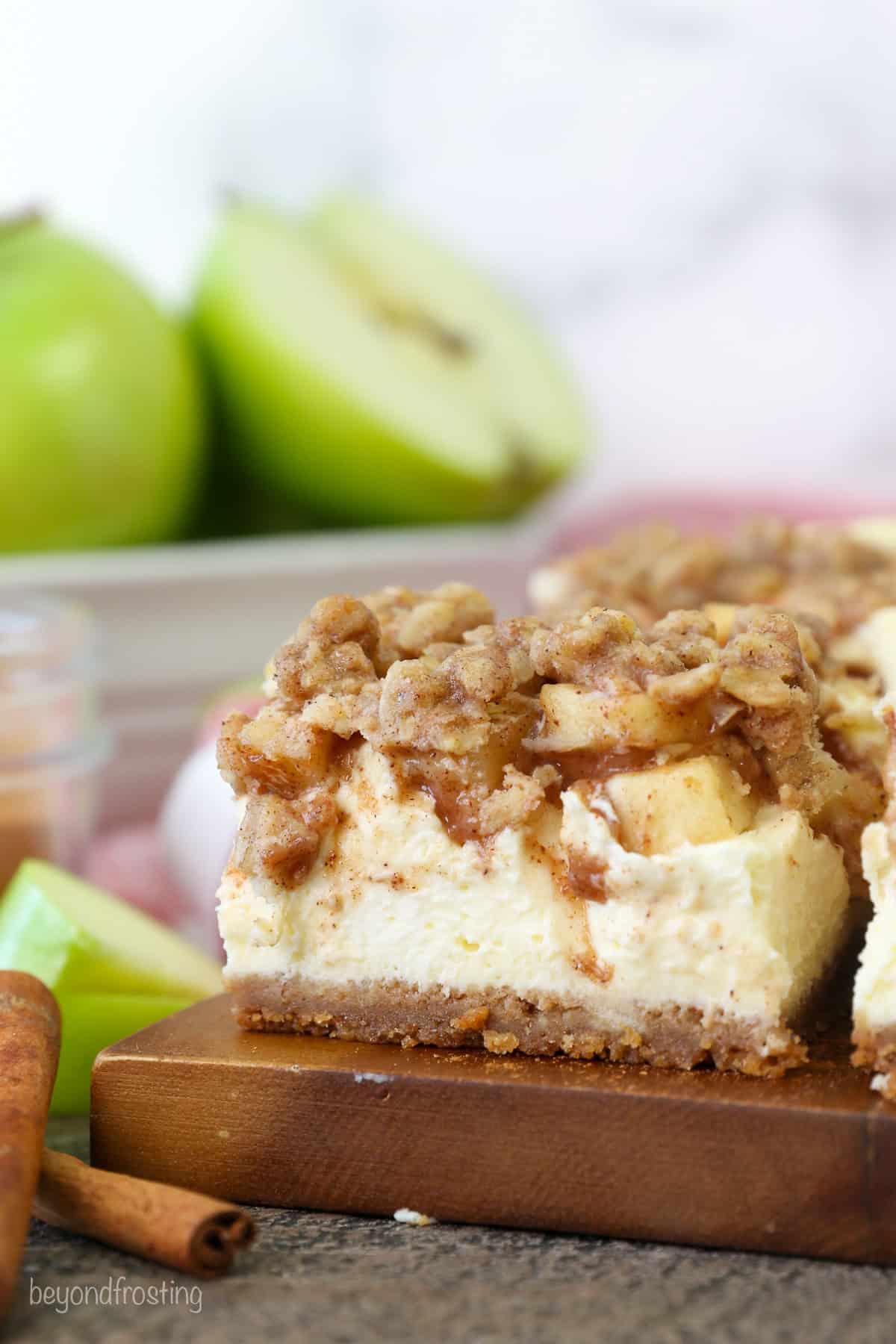 A caramel apple cheesecake bar on a wooden cutting board with a bowl of green apples in the background.